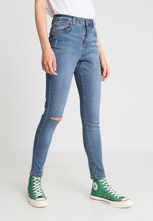 Persona che indossa jeans skinny azzurri chiari con uno strappo al ginocchio, maglietta bianca e sneakers alte verdi, in piedi su uno sfondo bianco.