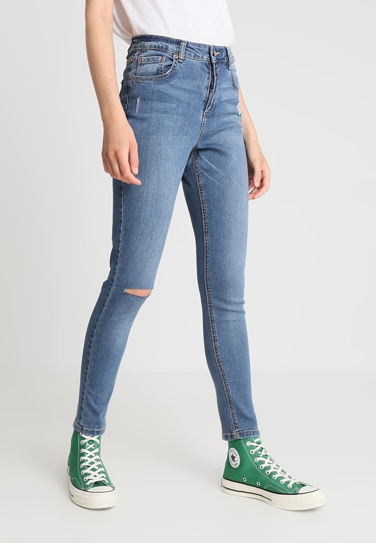 Personne portant un jean skinny bleu clair avec un trou au genou, un T-shirt blanc et des baskets montantes vertes, debout sur un fond blanc.