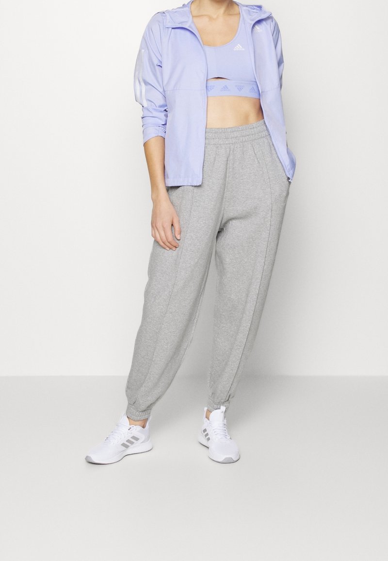 Femme portant une veste de sport légère mauve, un soutien-gorge de sport assorti, un pantalon de jogging gris et des baskets blanches, se tenant contre un fond blanc.