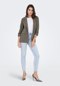 Blazer gris léger et texturé avec manches retroussées, associé à une chemise blanche ajustée et un jean skinny bleu clair. Talons neutres remarquables.