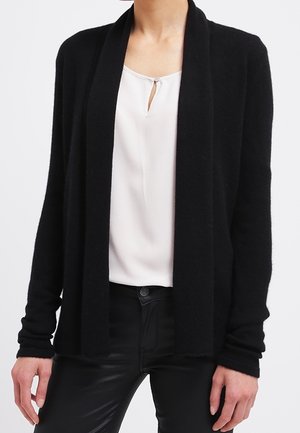 Vrouw draagt een zwarte open cardigan over een witte blouse met een keyhole-detail en zwarte broek, staand met ontspannen handen langs haar zij.