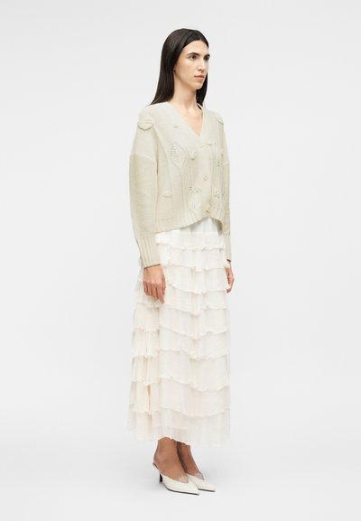 Γυναίκα με μακριά σκούρα μαλλιά που φοράει μπεζ πλεκτή ζακέτα, λευκή μακριά φούστα με στρώσεις βολάν και λευκά μυτερά παπούτσια με χαμηλό τακούνι, στέκεται πλάγια.