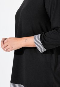 Maglia nera con maniche lunghe e polsini a righe in contrasto. Il materiale sembra morbido, testurizzato e leggero, adatto per un abbigliamento casual.