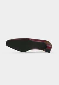 Chaussure de ville en cuir bordeaux, posée sur le côté, montrant une semelle noire avec un bout carré et un petit talon empilé.