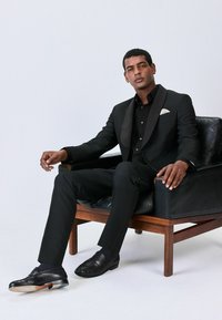 Musträga mustriga ülikond satiinist krae ja musta särkidega. Mustad läikivad loafer'id. Istub puidust käetugedega toolis.