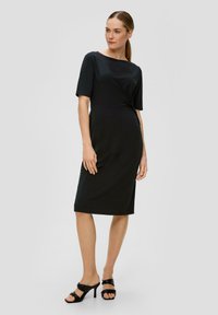 Robe midi noire à manches courtes, en tissu lisse, avec un détail torsadé à la taille, un col rond et un ourlet droit. Associée à des sandales noires à talons.