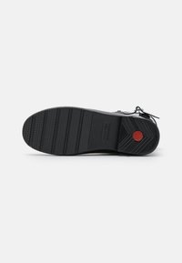 Semelle de chaussure noire avec une surface en caoutchouc texturé, présentant des rainures horizontales et un accent circulaire rouge au niveau du talon.