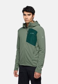 Giacca in pile verde con una tasca sul petto di un verde scuro a contrasto. Presenta un cappuccio, chiusure con zip e un tessuto elasticizzato per la mobilità.