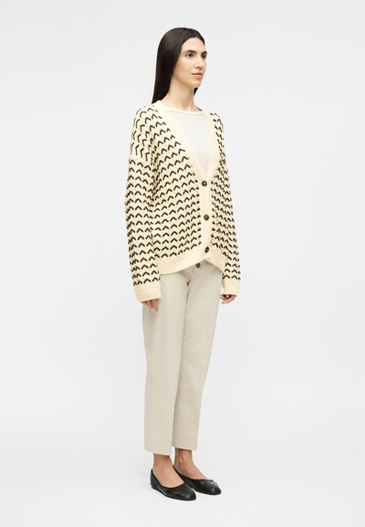 Γυναίκα που στέκεται φορώντας ένα εκρού πλεκτό ζακέτα με κουμπιά και σχέδιο, λευκή μπλούζα, μπεζ παντελόνι και μαύρα επίπεδα παπούτσια, σε απλό ανοιχτό φόντο.