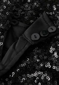 Tessuto nero tempestato di paillettes rotonde e riflettenti. Un morbido tessuto nero con tre bottoni è visibile sul bordo.