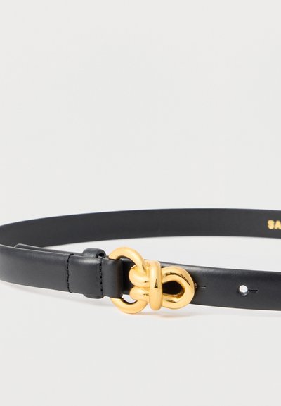 Ceinture en cuir noir avec une boucle nouée dorée brillante, montrant les détails de la couture et les trous de réglage sur un fond blanc.