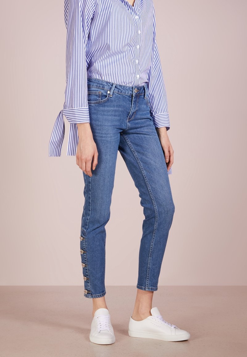 Vaqueros skinny de mezclilla azul con una cintura alta y botones laterales. Combinados con una camisa a rayas azul y blanca y zapatillas blancas.