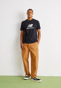 Black t-shirt with white "New Balance" logo, tan cargo pants, black and white sneakers. Simple design with a relaxed fit.