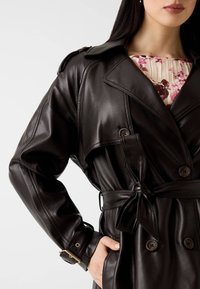 Brauner Leder-Trenchcoat mit doppelt geknöpftem Verschluss, weitem Kragen und verstellbarem Bindegürtel. Verfügt über eine Handtasche und eine glatte Textur.