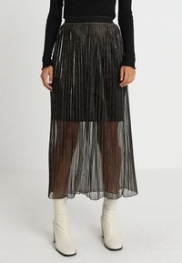 Jupe maxi plissée noire avec des fils métalliques, texture transparente, et une taille cintrée, portée avec des bottines blanches.