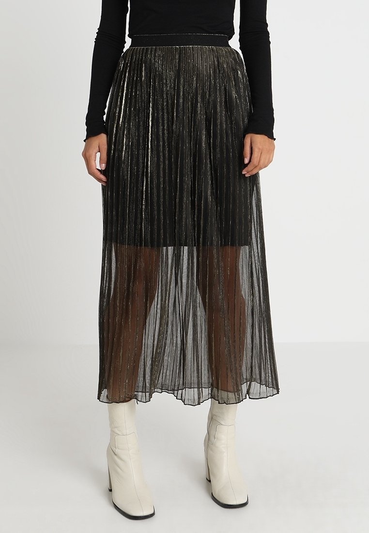 Jupe maxi plissée noire avec des fils métalliques, texture transparente, et une taille cintrée, portée avec des bottines blanches.