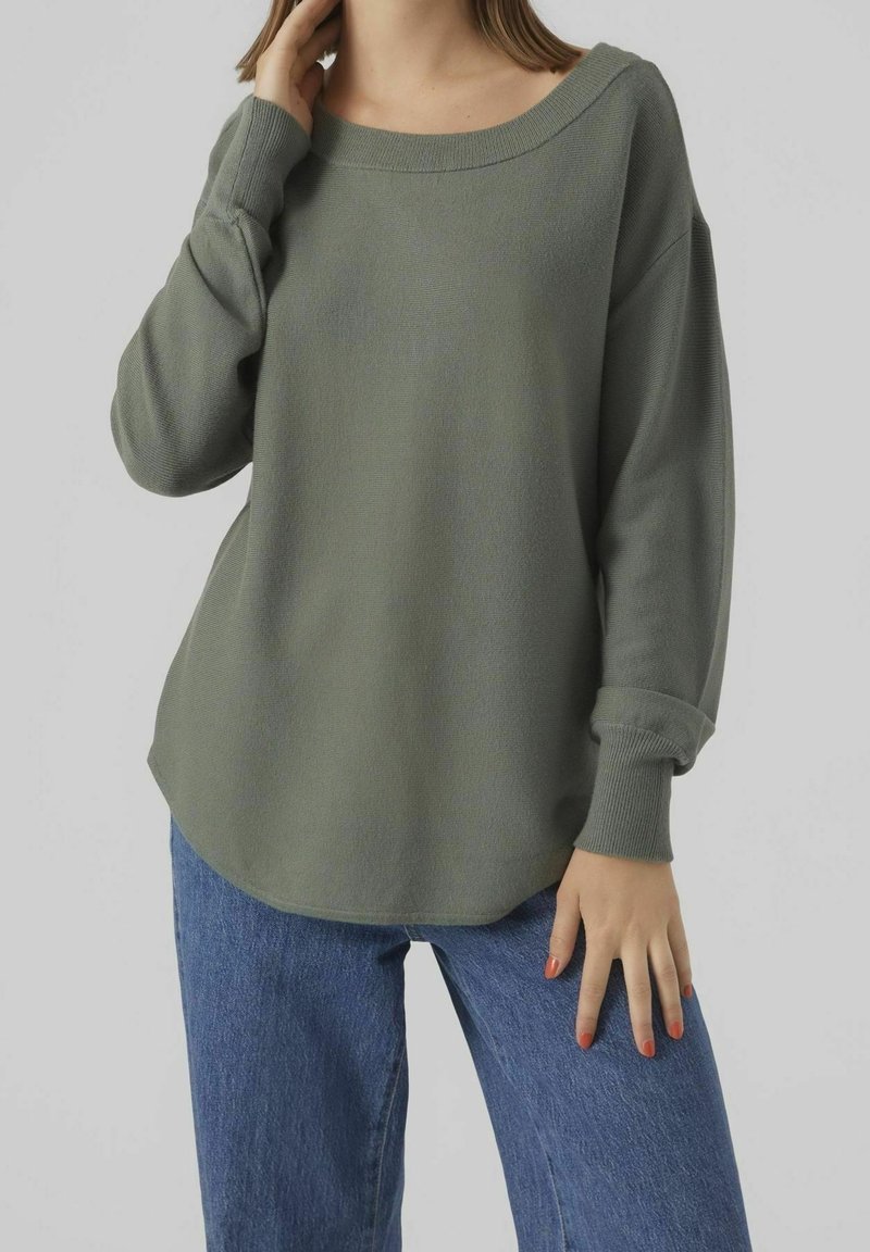 Femme portant un pull ample à manches longues couleur vert olive et un jean bleu, une main touchant son cou et l'autre reposant sur sa cuisse.