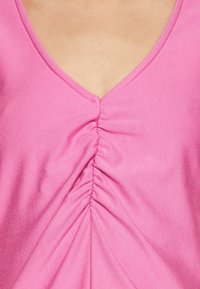 Růžový látkový top s hlubokým výstřihem do V a nařasením uprostřed, což vytváří texturovaný vzhled. Hladký, pružný materiál.