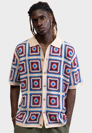 Młody mężczyzna z dredami nosi krótki rękaw, dzianinową koszulę zapinaną na guziki z czerwono-niebiesko-beżowym wzorem w kwadraty oraz oliwkowe spodnie.