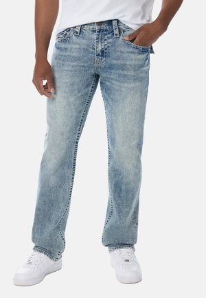 Mann trägt hellblaue ausgewaschene Jeans, weiße Sneaker und ein weißes Hemd, mit einer Hand in der Tasche, vor neutralem Hintergrund.