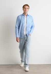 Camicia button-up azzurra, t-shirt bianca sotto, abbinata a jeans denim azzurri e sneakers bianche. Design semplice, vestibilità casual.