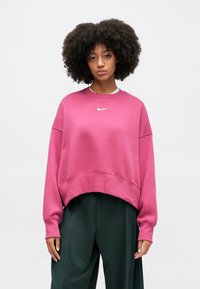 Rozā sporta krekls ar brīvu piegriezumu un ribotiem piedurkņu galiem. Uz krūtīm atrodas balts Nike logo. Apvienots ar tumši zaļām plisētām biksēm.
