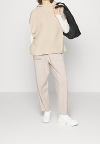 Top con cappuccio lavorato a maglia beige con bottoni laterali, abbinato a pantaloni larghi grigi chiari con texture. La modella indossa sneakers bianche e tiene una borsa nera.