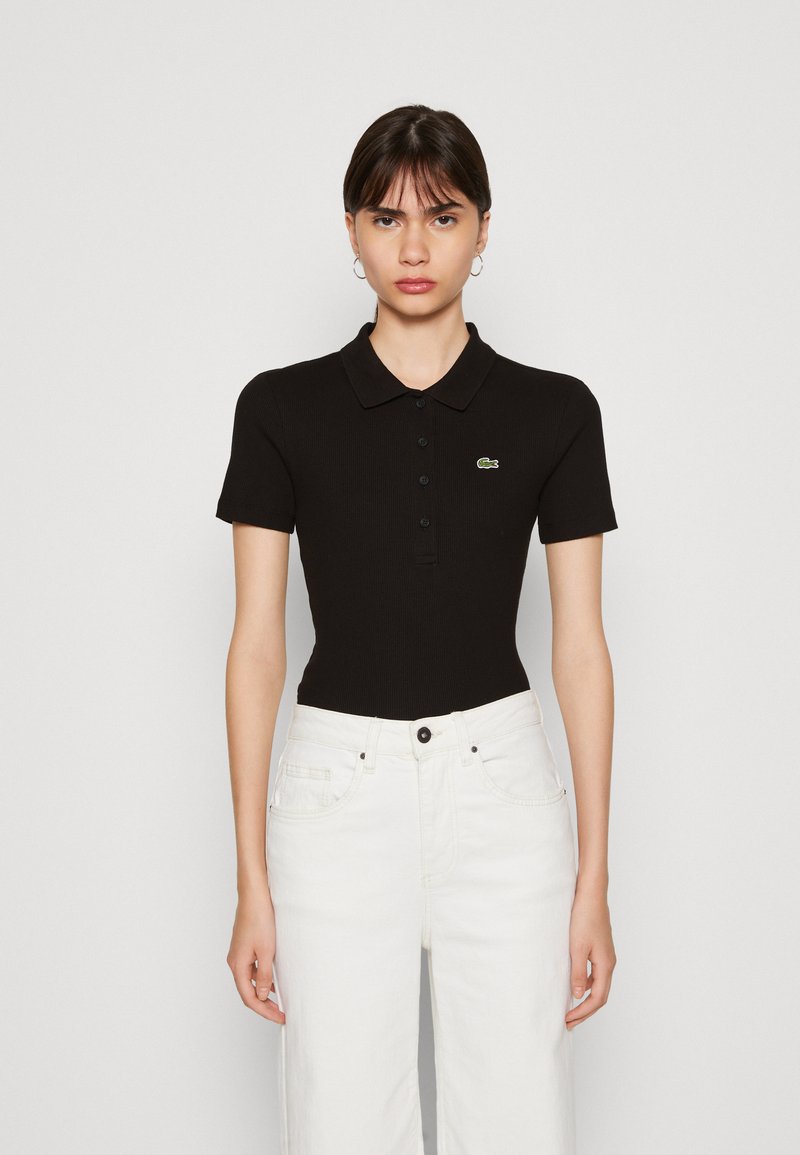 Polo shirt nera a righe con maniche corte, caratterizzata da un colletto e un logo verde Lacoste. Abbinata a jeans bianchi a vita alta.