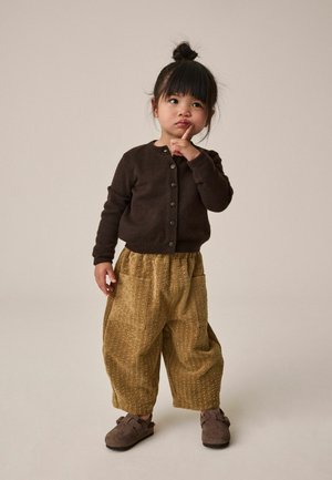 Junges Mädchen mit dunklen Haaren im Dutt, das eine braune Strickjacke, lockere senffarbene Hosen und braune Schuhe trägt und mit dem Finger auf den Lippen steht.