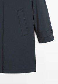 Ärmel und Seiten tasche eines dunkel marineblauen Mantels, mit einem Riemen mit Knopfverschluss am Ärmelbündchen, vor weißem Hintergrund gezeigt.