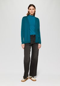 Tiefgrünes Langarmoberteil, unter einer offenen, dunklen Strickjacke geschichtet, kombiniert mit hochgeschnittenen schwarzen Jeans und beigen Plateauschuhen.