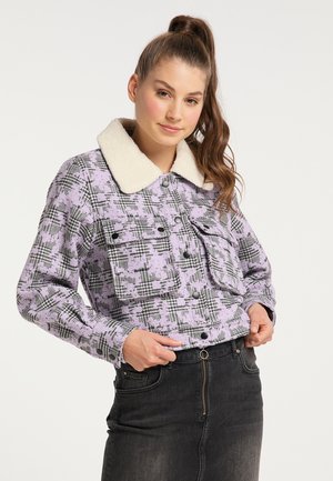 Junge Frau trägt eine violette karierte Jacke mit weißem Sherpa-Kragen und schwarzen Knöpfen, kombiniert mit einem dunklen Rock mit hohem Bund und Reißverschluss vorne.