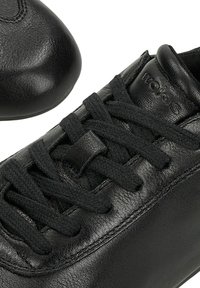 Schwarzer Ledersneaker mit schwarzen Schnürsenkeln, Nahaufnahme des Schnürbereichs und des auf der gepolsterten Zunge eingeprägten Logos.