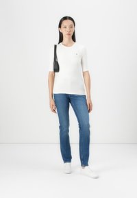 White short-sleeve top, fitted design, with a small logo on the chest. Paired with blue denim jeans and white sneakers.