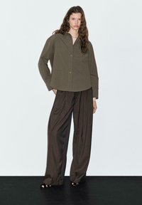 Olive green button-up shirt with a loose fit, paired with brown wide-leg trousers. Smooth fabric texture, minimal design, and no visible patterns.