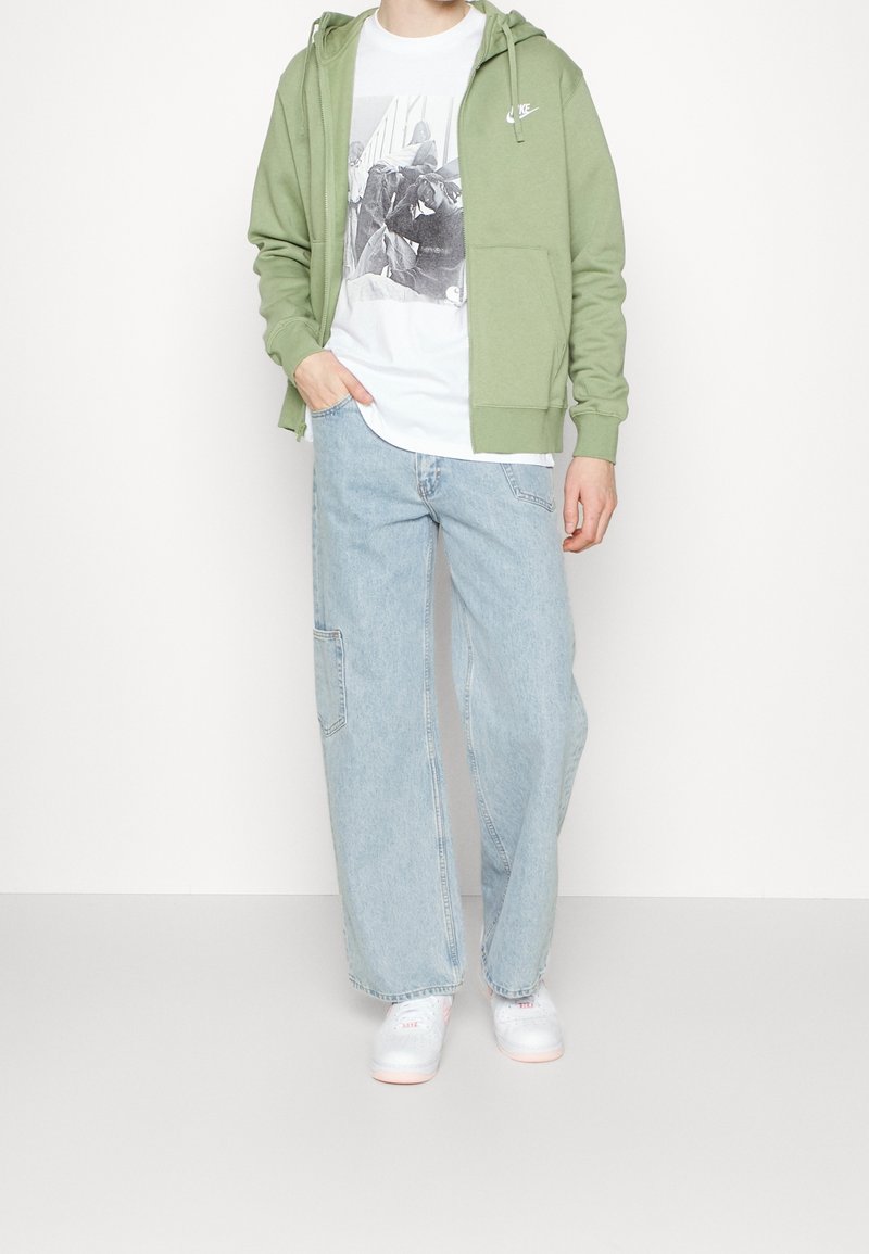 Personne portant un sweat à capuche Nike vert clair, un t-shirt blanc à motif graphique, un jean large bleu clair et des baskets blanches, debout avec une main dans la poche.