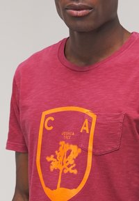 Mørkerød bomulds t-shirt med en forlomma, gul trægrafik og "JOSHUA TREE" tekst, med en afslappet rund halsudskæring.