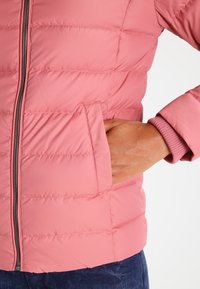 Main dans la poche d'une veste matelassée rose avec fermeture éclair, montrant le bord-côte du poignet et une partie d'un pantalon en denim bleu.