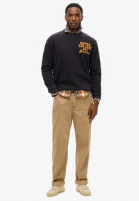 Sudadera azul marino con estampado de "SUPER COLLEGE", superpuesta sobre una camisa de cuadros. Combinada con chinos color beige y zapatillas de deporte en tono crema. Estilo sencillo y casual.