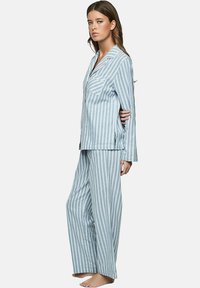 Lichtblauw gestreept pyjama set gemaakt van zachte stof. Beschikt over een geknoopte blouse met een zak en loszittende, rechte broek.