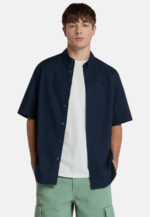 Kortærmet marineblå skjorte med knapper og krave. Indeunder ses en hvid t-shirt, og til det grønne cargo-shorts i lys farve. Enkel stil.