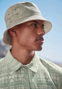 Giovane uomo con cappello a secchiello beige con motivo, piccolo orecchino a cerchio e camicia verde testurizzata con bottoni, che guarda a destra contro un cielo sereno.