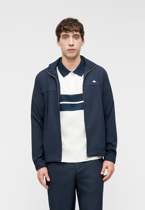 Jeune homme portant une veste zippée bleu marine sur un polo blanc à rayures bleu marine, et un pantalon assorti bleu marine, se tenant devant un fond uni.
