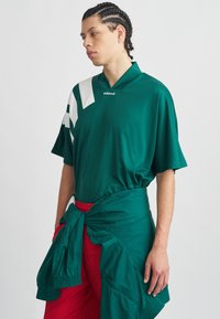 Jaunas vyras su pintomis plaukais, dėvintis žalią Adidas marškinėlį su baltomis juostelėmis, raudonas kelnes ir žalią striukę, susirištą aplink juosmenį.