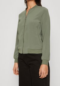 Olivgrön bomberjacka med dragkedja framtill, ribbade ärmslut och nederkant, med sidofickor och en slät, lätt glänsande textur.