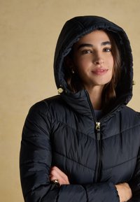Frau mit verschränkten Armen, die eine schwarze gesteppte Kapuzenjacke mit goldenem Reißverschluss und Kordelakzenten trägt, vor einem schlichten beigen Hintergrund.