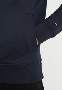 Felpa navy con polsini e orlo a costine. Presenta una tasca laterale e un piccolo dettaglio del logo colorato sulla manica. Texture liscia.