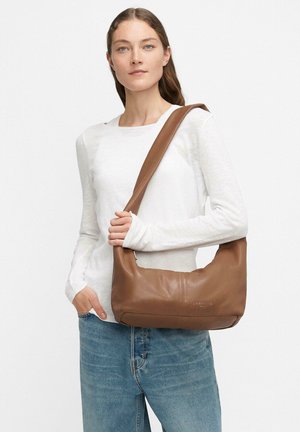 Donna con camicia bianca a maniche lunghe e jeans blu che tiene una borsa a tracolla marrone chiaro davanti a uno sfondo bianco semplice.