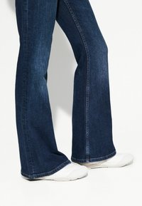 Dunkelblaue ausgestellte Jeans aus Denim, mit einem klassischen Fünf-Taschen-Design und einem Saum, der über weißen Sneakers liegt.