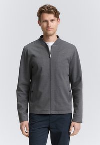 Szara kurtka na zamek z kołnierzykiem stójką, dwoma bocznymi kieszeniami z guzikami oraz gładką fakturą, zestawiona z białą koszulą pod spodem.